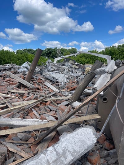 The image shows a scene of destruction in Terny, Ukraine, located at coordinates 50.979 latitude and 33.946 longitude. The debris of a building is scattered across a field, consisting of broken bricks, concrete slabs, twisted metal, and wooden beams. The remains of a large metal object, possibly part of a vehicle or machinery, are visible in the lower right corner. The environment is overgrown with trees and green vegetation. There are no people present in the image. The sky above is clear with white clouds against a blue background. The image suggests a past event of significant destruction, potentially caused by conflict or natural disaster.