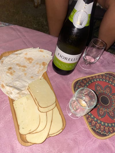 The image depicts a picnic scene, likely in Lagodekhi, Georgia, given the context provided. On a pink tablecloth, a wooden board holds several slices of light-yellow cheese and a stack of flatbread. To the right of the board, a bottle of Fiorelli Fragolino Bianco sparkling wine stands upright, with a few water droplets on its surface, indicating it's chilled. Next to the bottle, there are two clear glasses. One glass is empty and positioned to the right of the bottle. The second glass, closer to the foreground, contains a clear liquid, possibly water or more wine. A decorative coaster with a mandala-like pattern is placed beneath the second glass. In the background, parts of two individuals are visible, suggesting they are seated at the picnic. The ground appears to be grassy, with some dry leaves and twigs visible. The lighting suggests it is daytime, possibly late afternoon or evening.