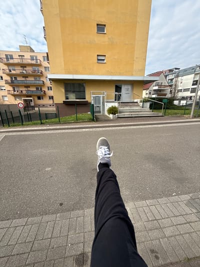 The image presents an outdoor urban scene in Lingolsheim, France, captured from a low, first-person perspective. In the immediate foreground, a person's left leg, clad in black trousers and a grey Adidas sneaker with white laces, is extended forward, occupying the lower right to mid-left portion of the frame.

An asphalt road, with a visible white lane marking, lies horizontally across the mid-ground. Adjacent to the road is a grey paved sidewalk. Beyond the sidewalk, a dark green metal mesh fence encloses a narrow grassy strip containing small white flowers.

The central background features a multi-story building with a light yellow facade. Its ground floor houses a commercial establishment accessible via a set of approximately five grey steps equipped with a silver handrail. The entrance consists of a glass door marked "NN." A large window to the left of the entrance displays the text "Cabinet ORTHODONTIE." Two small, rectangular windows are visible on the upper yellow facade.

To the left of the yellow building, a multi-story residential building with beige walls and dark-railed balconies is visible, topped by a television antenna. To the right, additional residential buildings of varying designs and colors are present in the distance. The sky above is partly cloudy with patches of blue visible. No other human figures are discernible in the scene.