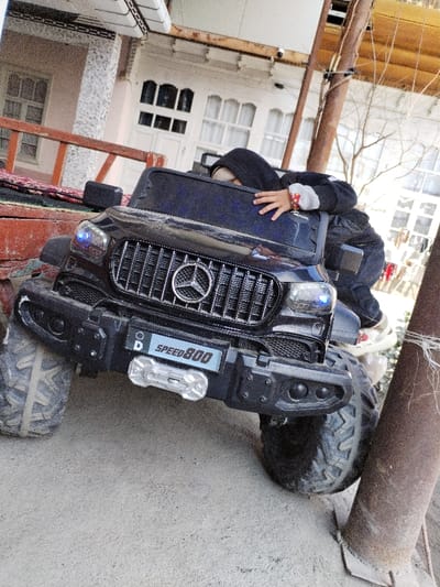 A single child is depicted, leaning over the dark-tinted windshield area of a black electric ride-on toy SUV, modeled after a Mercedes-Benz G-Class. The child wears a dark hooded garment and light-colored headwear, with their right hand visible on the toy vehicle's upper front section. Their lower legs and feet, clad in light-colored footwear with red accents, are partially visible near the right front tire. The toy SUV features a prominent Mercedes-Benz star logo on its vertical-slat grille, illuminated blue headlights, a robust front bumper, and a license plate displaying "D" and "SPEED 800" above a simulated silver winch. The vehicle rests on large, treaded tires on a light-colored, dusty ground. In the background, a light-colored building with white-framed windows and doors is visible, alongside a stone-clad pillar and a wooden railing partially obscured by a red patterned fabric. A dark brown utility pole stands on the right, with bare tree branches in the distant background. The scene is located outdoors in the city of Limbur, Uzbekistan.