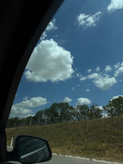 A car's side mirror reflects a cloudy blue sky and a grassy hillside with trees and a fence. The mirror itself is black and mounted on the car's frame, which is partially visible in the foreground. The road is paved and runs alongside the hillside. The scene appears to be captured from inside a moving vehicle, likely on a highway or a road outside of a city. The location is described as Poysdorf, Austria.