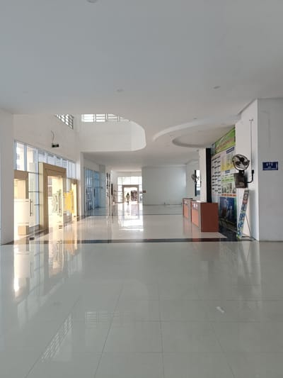 The image depicts an interior hallway of a modern building, likely a hospital or public institution, with highly reflective tiled flooring. The hallway is bright and spacious, with natural light entering through large windows on the left. White walls and a high ceiling with recessed lighting create an open and airy atmosphere.
On the left side of the hallway, there are several glass-paneled entrances with decorative elements. Further down the hallway, a person in a white garment is walking away from the camera, illuminated by light from the end of the corridor.
On the right side of the hallway, there is a reception or information desk area with two counter-like structures. Above the desk area, a large information board with text and images is mounted on the wall. Two oscillating fans are also attached to the wall. A blue sign indicating "UTD UNIT TRANSFUSI DARAH" is visible, suggesting a blood transfusion unit. A display stand with brochures is placed in front of the wall.
The overall impression is of a clean, well-maintained, and functional interior space. The presence of the blood transfusion unit signage suggests the location is a medical facility within Mesjid Reuntoh, Indonesia.