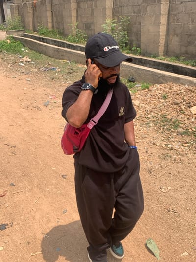 A male subject is depicted in an outdoor environment within Eika-Adagu New Layout, Nigeria, during daylight. He is viewed from a slightly elevated, oblique angle, oriented with his body facing right and head turned slightly left. The subject has dark skin and a beard, wearing a black baseball cap with a white "Champion" logo, a dark-colored ribbed short-sleeved collared shirt with a gold "RK KIEBAK" emblem on the left chest, and loose dark-colored trousers. He wears dark sneakers with white soles and light blue/green accents. A black digital smartwatch is present on his left wrist, and a red/pink crossbody bag with a wide strap is worn across his chest. The subject holds a dark-colored smartphone to his right ear with his right hand, indicating he is engaged in a phone call. His left hand is partially visible, bearing a blue wristband, resting at his side or in a pocket. The ground consists of unpaved light brown soil with scattered small stones and sparse debris, including a prominent green leaf near his right foot. In the background, a low concrete block wall is visible, behind which sparse green vegetation grows. Parallel to the wall, two linear, open concrete drainage channels are present. Bright sunlight casts a distinct shadow of the subject to his left on the ground.