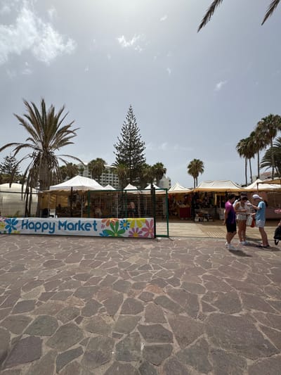 The image shows a street market in Playa de Las Américas, Spain. The market is set up with several stalls under white and tan tents, selling various goods. A banner in front of the stalls reads "Happy Market" and is decorated with colorful flowers. The ground in front of the market is paved with irregular-shaped stones. In the background, there are palm trees and a large building. Several people are browsing the stalls, and some are interacting with each other. The sky is cloudy.