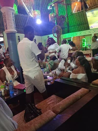 The scene is an open-air bar or restaurant with a thatched roof and wooden beams. The bar is located in Diani Beach, Kenya at the coordinates provided (latitude: -4.276, longitude: 39.592). There are several people seated at tables, some of whom are drinking from glasses. A woman in a white dress is standing at a table and speaking into a microphone. A man in a white shirt is standing next to her and looking at his phone. A few people are seated on large brown cushioned booths that line one wall. A black purse is sitting on the booth in the foreground of the image. A man in a white shirt is standing in the middle of the image. There are several drinks and bottles of alcohol on the tables. The environment is festive and lively. A television screen is visible on the wall, displaying footage of a sports game. There are a few potted plants, a large fan, and a wooden wheel hanging from the ceiling. There is a bar area behind the people, where there are bottles of liquor and a cash register. The scene appears to be taking place at night or in the evening, as the lighting is low and there are a few spotlights shining on the tables.