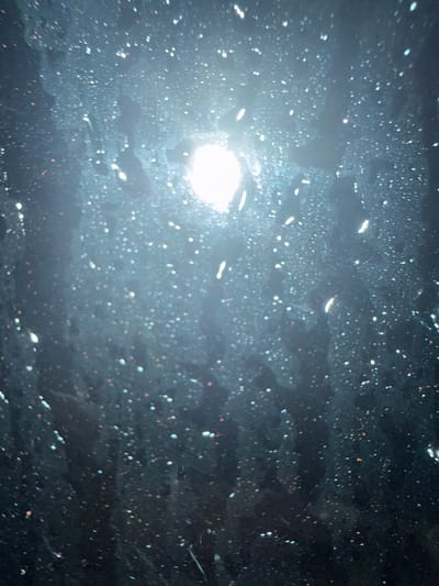 The image shows a close-up, abstract view of a surface covered in water droplets, possibly a window or a windshield, illuminated by a bright light source. The background is dark, and the water droplets create a speckled effect, with some appearing as white highlights and others reflecting light in various colors, including hints of orange and blue. The light source is off-center and appears to be a bright, possibly blown-out, bulb or the sun. The overall impression is one of looking through a wet surface at a light. There is no discernible location in Long Thành, Vietnam, visible in this abstract image.