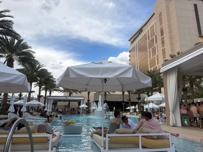 This image shows a vibrant pool scene in Las Vegas, United States of America. The "Durango" hotel towers in the background, suggesting the location is at or near the Durango Casino & Resort. In the foreground, several people are lounging on poolside beds under white umbrellas. A pool stretches across the middle of the scene, with a fountain visible in its center. People are swimming, sunbathing, and interacting in and around the pool. Palm trees line the perimeter of the pool area, adding to the resort atmosphere. A bar area with patrons seated at the counter can be seen to the right of the pool. The sky is partly cloudy.