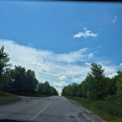 The image depicts a paved road winding through a forested area, likely in the city of Fursy, Ukraine. The road surface is worn and shows signs of disrepair, with patches of gravel and potholes. Green trees line both sides of the road, forming a dense canopy that extends into the distance. The sky above is a bright blue, with scattered white clouds, some of which are wispy and streak across the sky. The scene appears to be captured from a moving vehicle, as suggested by the perspective and the presence of what appears to be the top edge of a windshield. There are no people or animals visible in the image. The overall impression is one of a rural, somewhat neglected, but scenic road under a clear sky.