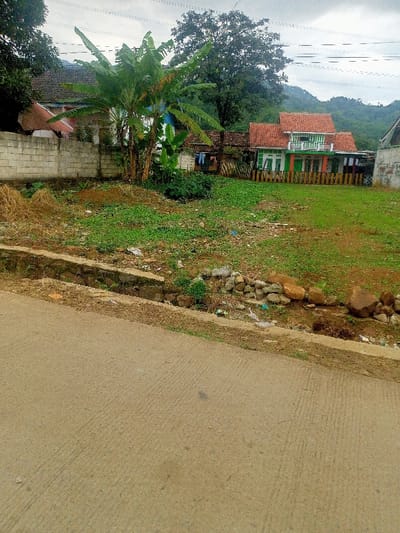 The image captures a rural or semi-rural landscape in Cimaung, Indonesia, under an overcast sky during daytime. In the immediate foreground, a textured, light-colored paved road occupies the bottom portion of the frame. Adjacent to the road is a shallow concrete and stone drainage ditch, which appears dry or with minimal water.

Beyond the ditch lies an open, somewhat unkempt plot of land. This area is covered with patchy green grass, various weeds, and visible dirt, with scattered rocks and some dry debris. A prominent cluster of banana trees stands in the midground of this grassy lot, their large leaves fanning out.

Behind the open plot, several houses are visible. On the left, a house is partially obscured by a light-colored concrete block wall. In the center-right, a more distinct house features a characteristic reddish-orange tiled roof, green and white painted walls, and a small balcony. A colorful wooden fence, painted in alternating orange and green segments, runs along the front of this property. Clothes are seen hanging on a line near one of the houses.

In the background, lush green hills or mountains rise under the cloudy sky, extending across the horizon. Power lines stretch across the upper part of the sky. The scene is devoid of people and depicts no specific activity, offering a tranquil glimpse into a residential area integrated with natural surroundings. No visible text is present.