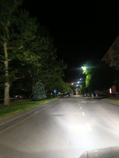The image depicts a nighttime scene on an asphalt road in Pivdenne, Ukraine, characterized by street lighting and surrounding greenery. On the left side of the road, lush trees line a grassy area, with one distinct conical evergreen tree visible. Further left, a blue parking sign with a white "P" is mounted on a pole. The road itself is marked with white dashed lines. Streetlights illuminate the scene from the right side of the road, casting a bright glow. To the right, a multi-story building with visible windows is situated. The perspective suggests the image was taken from a moving vehicle, as indicated by the blurred motion of the trees and the bright headlights illuminating the road ahead. There are no people or animals clearly visible in the frame.