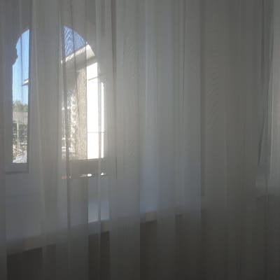 A semi-transparent white curtain hangs in front of a window, partially obscuring the view of a room and the outdoors. The window itself appears to be an arched window with a grid pattern, and light is streaming through it, creating a bright, diffused glow. Through the curtain, one can faintly discern the outlines of buildings and trees outside, suggesting a residential or semi-rural setting. The curtain is layered, with some panels appearing thicker and more opaque than others, creating variations in light and shadow. The overall impression is one of soft, diffused light and a sense of privacy. The specific location in Cepeleuți, Moldova, cannot be determined from the visual information provided.