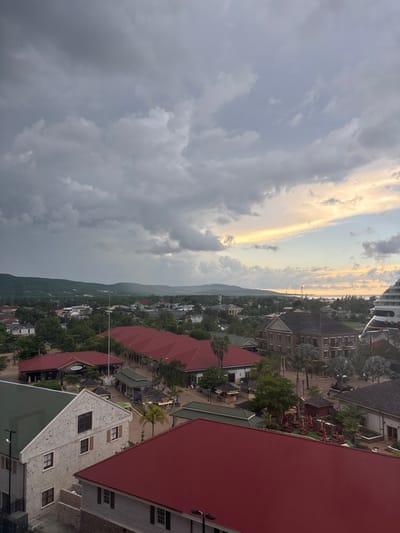 This is an aerial view of Falmouth, Jamaica, under an overcast sky transitioning from dark, heavy clouds in the upper left to a lighter horizon with hints of yellow and orange sunlight on the right.

The immediate foreground features the tops of various buildings. A prominent light-colored building with a grey gabled roof and several windows with dark shutters occupies the bottom-left, partially cropped. Adjacent to it, a large structure with a red corrugated metal roof is visible.

In the midground, a significant portion of the city's commercial or port district is visible. A series of connected buildings with uniform red corrugated metal roofs stretch horizontally across the frame, forming what appears to be a marketplace or shopping area. Numerous smaller kiosks or stalls with dark or green roofs are arranged in open pedestrian spaces among these buildings. Palm trees and other tropical foliage are interspersed throughout this area. To the right of this complex, a substantial multi-story building constructed of dark-colored brick or stone with white trim and a dark roof stands out. Further to the right, the upper decks and white hull of a large cruise ship are partially visible, indicating a docking area.

The background shows the town extending inland, with a mosaic of residential and commercial structures. Green, forested hills and mountains rise behind the town, extending to the distant horizon. A distinct light-colored building with a tall steeple or tower is discernible in the mid-left background. The sea is visible on the far right horizon, reflecting the lighter sky conditions. No specific human figures or explicit actions are discernible due to the elevated perspective and distance, but the urban infrastructure implies human presence and activity.