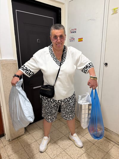 A woman stands in a doorway in Ashkelon, Israel, holding two shopping bags. She is wearing a white patterned top and shorts with a black and white pattern, white sneakers, and glasses. A black crossbody bag is slung over her shoulder. The doorway appears to be in an apartment building, with tiled flooring and a metal door. There are stickers on the wall to the right of the doorway. The woman is smiling and sticking her tongue out. The shopping bags contain groceries and other items.