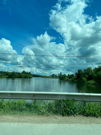The image shows a view from a vehicle traveling along a road next to a body of water, likely a river or lake. A metal guardrail is positioned between the road and the water. Lush green vegetation grows along the edge of the water. The water surface reflects the sky and the surrounding trees. Power lines are visible against the backdrop of a cloudy blue sky. The image was taken near the city of Норья, Russia. The scene depicts a natural environment adjacent to an infrastructure element, suggesting a rural or suburban location.