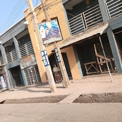 This outdoor daytime image, likely taken in Dakata, Nigeria, presents a tilted perspective of a street lined with buildings and various elements of urban infrastructure under clear, sunny skies.
The scene features a row of two-story buildings, alternating in color between yellow-orange and blue-grey, each equipped with balconies featuring white horizontal railings on the upper floor. In the foreground and midground, two prominent utility poles stand. The pole on the right side of the image has the word "POSS" painted vertically in blue capital letters. Attached to this pole is a political campaign poster depicting two men, one dressed in white and the other in blue, with visible text such as "NUHU SARA," "DARA," "SALLAM," and "SAMUSA."
Near the "POSS" pole, a person is partially visible, dressed in a white head covering and a long, patterned dress. To the right, an open storefront with a corrugated metal awning reveals a collection of wooden furniture or structures inside, possibly a stall or large seating. Another person is partially visible on the far left, near a shop with a metal security gate.
The street itself is visible in the lower foreground, bordered by a concrete sidewalk or curb. A wooden plank bridges a gap in the curb, seemingly providing access across a small ditch or drain. The bright sunlight casts distinct shadows from the poles and buildings, indicating the time of day. The presence of shops, political posters, and utility services like "POSS" suggests a commercial or mixed-use area.