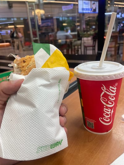 A person's hand is holding a Subway sandwich wrapped in a white paper napkin. To the right of the sandwich is a red Coca-Cola cup with a white lid and straw. The scene appears to be indoors, possibly a food court or restaurant, with blurred background elements suggesting a public space with seating and displays. The image quality is good, and the focus is on the food and drink. The presence of a Subway and Coca-Cola branding suggests a typical fast-food dining experience. There is no information to suggest this location is in Gollapalle Khurd, India.