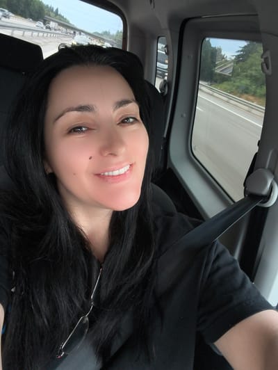 A woman with long, black hair and a mole on her cheek is taking a selfie in a car. She is wearing a black t-shirt and has sunglasses hanging from her hair. The car appears to be on a highway. The background shows a blurred view of the road, trees, and other vehicles. There is no indication that this image was taken in Baden-Baden, Germany.