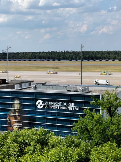 This is a medium shot of Albrecht Dürer Airport Nuremberg, Germany, on a partly cloudy day. The airport terminal is a modern building with a blue glass facade. A large advertisement featuring a portrait of Albrecht Dürer is displayed on the facade, alongside the airport's logo and name. In the background, the airport tarmac is visible, with several yellow vehicles, a white truck, and airport infrastructure. A line of solar panels is visible in the distance, beyond the tarmac and a dense forest. The foreground is dominated by lush green trees and bushes.