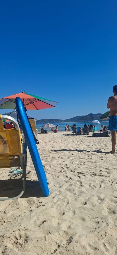 The image depicts a beach scene in Armação Dos Búzios, Brazil, observed in daylight under a clear blue sky. The foreground features beach paraphernalia including a multi-colored sun umbrella with alternating panels of red, green, and orange, as well as a blue object, possibly a surfboard or beach lounger, resting on a yellow beach chair with a gray frame.
Further into the scene, a crowd of people are present on the sand, some appearing to be sunbathing or relaxing under various beach umbrellas. The sea can be seen with landmasses visible in the distance. At the very top edge of the image, the upper torso of a person with blue swimwear is slightly visible, situated closer to the top right corner of the frame.
The sand in the foreground appears light in color with visible grains and textures. The overall perspective suggests the photo was taken either from a standing position or from a slightly elevated point on the beach.