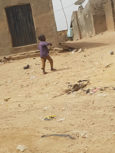 A daytime shot shows a child walking barefoot on a dirt ground in Dutsinma B, Nigeria. Litter is scattered on the ground. The child is wearing a purple shirt and brown pants. To the left is an old building with closed, barred windows. A satellite dish is mounted on the building's roof.