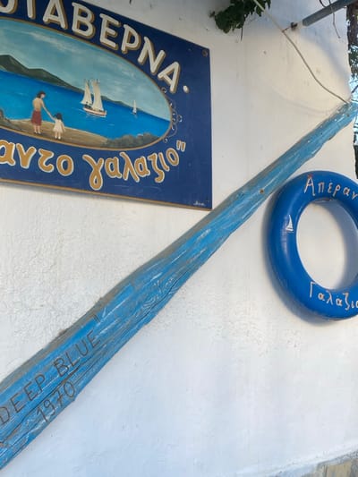 The image depicts the exterior of a building, likely a taverna, in the city of Απολλωνία, Greece. A prominent dark blue rectangular sign with a painted oval scene is mounted on a textured, whitewashed wall, positioned towards the upper left. The sign's scene portrays a woman and a small child on a sandy beach, facing a calm blue sea where two white-sailed boats are visible with distant land in the background. Above the oval, the Greek word "TAVERNA" is written in white capital letters. Below the oval, the Greek phrase "ΑΠΕΡΑΝΤΟ ΓΑΛΑΖΙΟ" (Endless Blue) is written in gold.
Below and to the right of the sign, a long, slender wooden object, painted a distressed light blue, extends diagonally across the mid-ground from the lower left towards the upper right. Carved or painted into this object are the words "DEEP BLUE" and "1970." To the right of this wooden object and centered vertically, a blue lifebuoy or ring is affixed to the white wall. This lifebuoy features the Greek words "Απέραντο" at the top and "Γαλάζιο" at the bottom, written in a light color, and includes a small white sailboat icon on its inner edge.
The whitewashed wall shows minor textural variations and some shadows from the objects and an overhead structure or vine, part of which is visible in the upper right corner. White electrical wiring is also present near the top of the wall. The very bottom edge of the image reveals a darker, stone or concrete base structure.