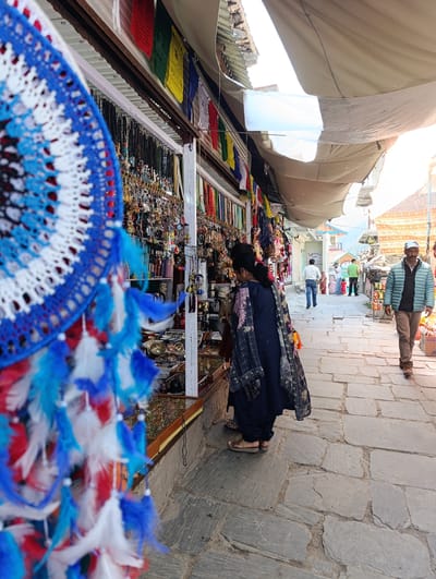 In a narrow, stone-paved alleyway lined with market stalls, a woman in a dark blue garment and patterned scarf is browsing. She is facing a stall displaying a wide assortment of necklaces, keychains, and other trinkets. In the foreground, a large, colorful dreamcatcher with blue and white feathers hangs, partially obscuring the view. Beyond the woman and the stall, the alley continues, with more people walking and other shops visible. Prayer flags are strung along the roofline of one of the stalls. The scene is outdoors, with natural light filtering through an overhead canopy of fabric. The overall impression is of a bustling marketplace.