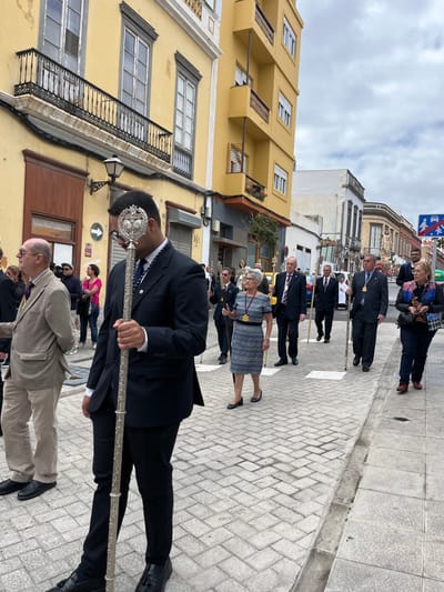 The image shows a procession of people walking down a street in the city of Las Palmas de Gran Canaria, Spain. The street is paved with grey stone, and there are yellow buildings on either side. The people are mostly wearing formal attire, and some of them are holding staffs. The man in the foreground is wearing a dark suit and holding a silver staff. The street is mostly empty, with only a few other people visible in the background. The image was likely taken on a sunny day, as the sky is clear and the sun is shining on the buildings. The coordinates (latitude: 28.099, longitude: -15.418) place the scene on the street Calle de Triana, near the Plaza de San Antonio Abad in the Vegueta neighborhood of the city.
