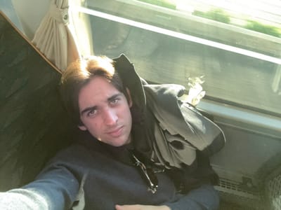 A single male individual, appearing to be an adult, is depicted in a selfie-style photograph taken within a vehicle, specified as being in the city of Ladispoli, Italy. The male has medium-length dark hair and is looking directly into the camera with a neutral expression. He is wearing a dark blue long-sleeved sweater. A dark, possibly black, hooded garment is draped over his left shoulder and arm. Black-framed sunglasses are tucked into the front neckline of his sweater.

The background shows the interior of a moving vehicle, likely a train or bus. To the upper right, a large window reveals a blurred green landscape, indicating motion and an outdoor environment. Strong natural light emanates from this window, illuminating the right side of the individual's face and hair. Below the window, a light-colored horizontal panel is visible, with a section of horizontal grilles further to the right, possibly an air vent or part of the vehicle's interior structure. To the left of the individual, a dark, textured surface, likely upholstery or a wall panel, is present. Above the individual's head, a light-colored fabric curtain, partially secured by a metal eyelet, is visible near the upper left corner of the frame.