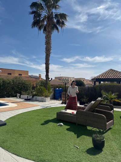 A woman is observed standing outdoors in a residential property in Fuente Álamo de Murcia, Spain, under a clear blue sky with sparse cirrus clouds. She wears a long-sleeved white top and a long, striped red and white skirt. Her dark hair is pulled back. She holds a small, light-colored object in her right hand and another in her left hand, which is slightly obscured.
The scene features a green artificial lawn in the foreground. To the left, a swimming pool with blue tiles is partially visible, surrounded by beige and grey patterned paving. To the right of the woman are two dark brown wicker lounge chairs, one of which has a red object (possibly a phone) and a silver laptop on its cushion. A pair of white slippers rests on the grass near the chairs. Further right, a dark potted plant is visible.
In the midground, a tall palm tree with a thick, textured trunk stands prominently. Behind the woman and the lounge chairs, a low wall separates the lawn from a paved area with various plants, a large golden-colored head sculpture, and two blue plastic barrels stacked vertically. A decorative black lantern post with three lights is positioned further back. In the background, several Mediterranean-style houses with light-colored walls and tiled roofs are visible, interspersed with trees and shrubs. One rooftop displays solar panels. A brown-tiled gazebo structure is visible on the far right of the background.