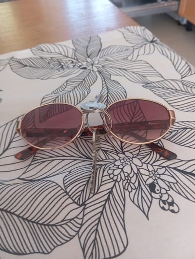 A close-up, high-angle shot shows a pair of gold-rimmed sunglasses with reddish-brown lenses resting on a white cloth with a black line drawing of stylized leaves and flowers. A silver key is attached to the sunglasses' temple and hangs down between the lenses. The sunglasses have a tortoiseshell pattern on the arms. In the background, the edge of a wooden table is visible, and beyond that, a blurred view of a room with furniture. The scene appears to be indoors, possibly in a home or a café in Benissa, Spain, given the context of the prompt. There are no people present.