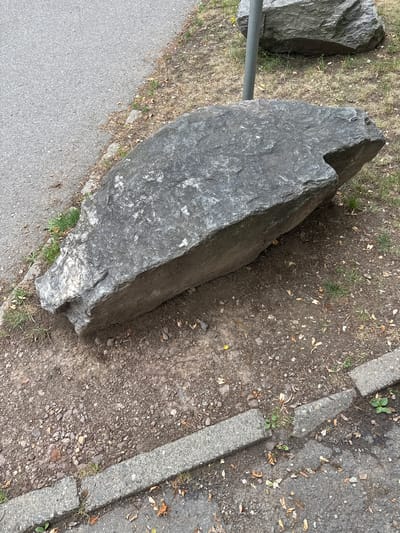 This media file is a horizontal, eye-level shot of a large, rough, gray boulder resting on a patch of dirt and dry grass next to a paved surface. The boulder takes up a significant portion of the frame, with its angular, weathered texture clearly visible. A thin, horizontal metal pole extends from the left side of the frame, passing in front of another, smaller boulder on the left. The ground next to the boulder is a mix of dirt, small stones, and scattered dry leaves.
To the right of the boulder, there is a concrete curb with some growth of green grass poking through, separating the dirt patch from an asphalt surface. The asphalt appears to be a road or pathway. The lighting suggests it is daytime, possibly a sunny day given the shadows cast by the boulder.
There is no discernible activity or event taking place; the scene appears static, focusing on the natural and man-made elements of the environment. The location is identified as Chemnitz, Germany. No text is visible in the image.