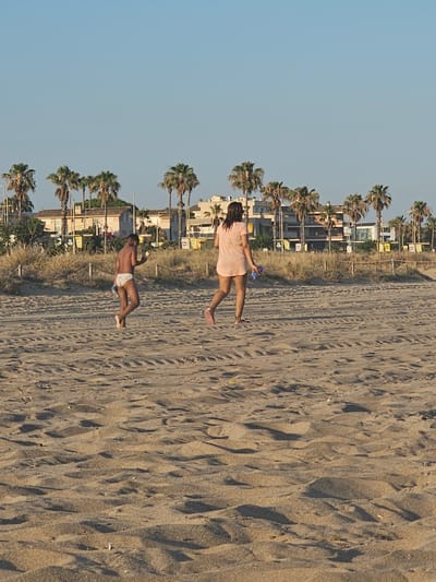 In Castelldefels, Spain, two individuals are walking on a sandy beach. In the foreground, the sand is textured with tire tracks and natural undulations. In the midground, a young person wearing white patterned swim trunks walks towards the left, holding a blue object in their right hand and a light-colored item in their left. To their right and slightly further back, a person wearing a short, pale pink dress walks towards the right, carrying a blue object in their left hand. In the background, a line of palm trees is visible, with buildings and some vegetation behind them. The sky above is clear and blue.