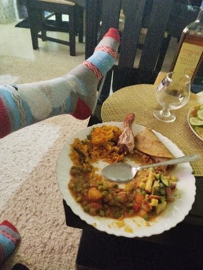 A white plate holds a partially eaten meal of rice, a chicken drumstick, a quesadilla, vegetables, and a stew. A silver spoon rests on the plate. A glass and a bottle of whiskey sit on a golden placemat. A foot clad in a red, grey, and blue sock is casually propped up near the plate. The setting suggests a relaxed, informal dining experience.