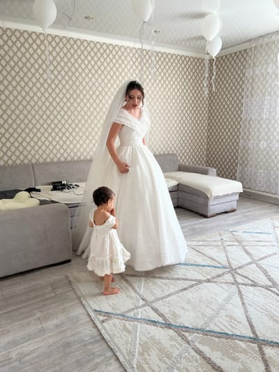 A woman in a sparkling white wedding dress and veil stands in a room, looking down at a young child in a white dress. The woman's hair is pulled back, and she is wearing a necklace. The child is facing away from the camera, with their hands clasped in front of their mouth. In the background, a gray sectional sofa is visible, with some objects on it. The walls are covered in patterned wallpaper. There are white balloons tied to the ceiling with ribbons. A patterned rug is on the floor. The lighting suggests an indoor setting. The location is identified as Astrakhanka, Kazakhstan.