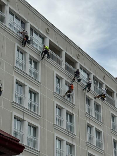 In Kislovodskiy, Russia, a group of individuals are engaged in high-rise window cleaning on the exterior of a multi-story building. The building features a light-colored facade with white balconies and numerous windows. The individuals are suspended by ropes attached to the top of the building, employing specialized equipment for their work. They are equipped with safety harnesses, ropes, and buckets containing cleaning supplies. The workers are positioned at various levels along the building's facade, indicating a systematic approach to cleaning. The sky is overcast, suggesting a potentially cloudy day. The image captures the challenging nature of this profession, highlighting the skill and courage required for such tasks.