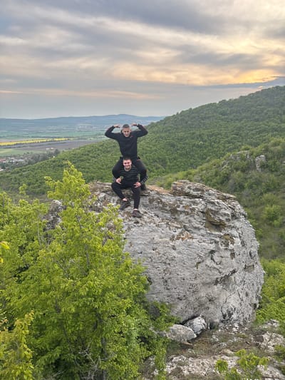 Two men are posing on a large rock outcrop. The man on the left is standing and flexing his arms. The man on the right is lying on his side. Both are wearing dark clothing. The rock is light gray with some darker patches. The background shows a mountainous, forested area with a valley. There is a small village in the valley. The sky is cloudy with a hint of sunset. This is in Goritsa, Bulgaria.