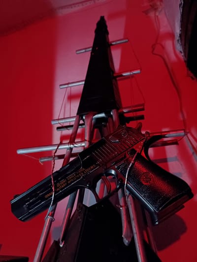 A black Desert Eagle pistol is propped on a metal stand with red lighting. The pistol has the markings "DESERT EAGLE PISTOL" and "ISRAEL MILITARY INDUSTRIES LTD (I.M.I.)". The background is a solid red wall. The scene appears to be indoors, with no visible indicators of the city of Tash-Dobo, Kyrgyzstan.