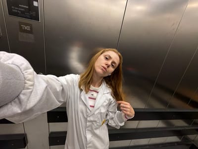 A young woman with blonde hair, wearing a white lab coat over a striped shirt, stands in an elevator. She is taking a selfie with her left arm extended. The elevator walls are made of brushed metal, and there is a sign visible on the left wall that reads "evolution 40 personen 3000 kg" and the TKE logo. The scene appears to be indoors, possibly in a modern building. The lighting is artificial and somewhat diffused.