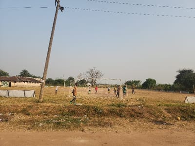 The image captures an outdoor scene in Umuoye, Nigeria, depicting several children playing football on a dry, unpaved field under a clear, bright sky.
In the foreground and prominent on the left, a tall wooden utility pole with overhead wires extends upwards. The field itself is characterized by its brownish-orange, dry soil, with patches of sparse vegetation. Towards the middle ground, a makeshift football goal with a net is set up, around which a group of children are actively engaged in play. Some children are clustered near the goal, while others are spread out across the field, indicating an ongoing game. One child in a brown top is particularly visible in motion near the left side of the goal. On the far right foreground, a yellowish ball lies on the ground.
In the background to the left, a long, single-story building with a light-colored, possibly painted, facade and a dark roof is visible, suggesting a school or institutional structure. This building is partially obscured by trees and other foliage, including a few palm trees. On the right side of the field, further white rectangular structures, possibly temporary barriers or markers, are present.
The clear sky and distinct shadows indicate it is daytime, likely mid-morning or late afternoon, with sunny and dry weather conditions. The overall scene suggests a casual recreational activity in a rural or semi-urban environment. No discernible text is visible in the image.