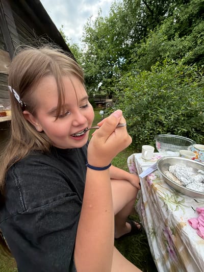This media file shows a young girl eating from a spoon outdoors in a backyard setting in Бронница, Russia. The girl is lying down, with her head tilted upwards and her eyes closed as she enjoys her food. She is wearing a black t-shirt and dark jeans. Her hair is long and light brown, held back by a white clip. A dark blue bracelet is visible on her wrist.

In the background, a picnic table is set with a floral tablecloth. On the table are various items, including a clear bowl, a metal bowl covered in aluminum foil, a few white cups, and some pink napkins. To the left of the girl, there is lush green foliage and a cloudy sky can be seen. The overall impression is of a casual outdoor meal on a possibly warm day, with a focus on the girl's enjoyment of her food. No visible text is present in the image.