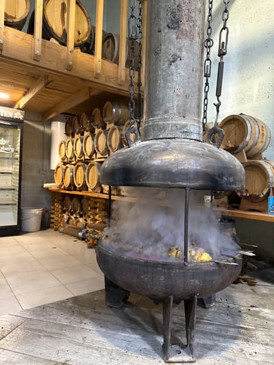 In Sisian, Armenia, within what appears to be a brewery or wine cellar, a large, rustic metal brazier is actively smoking, with several skewers of meat cooking over an open flame. The brazier is suspended from a heavy metal chain mechanism and features a wide, dome-like cover that directs the smoke upwards through a tall metal chimney. The scene is set against a backdrop of stacked wooden barrels, suggesting an environment where aging beverages takes place. To the left, a commercial refrigerator unit, possibly for drinks, is visible. The floor is tiled in the foreground and transitions to weathered wood planks. The overall atmosphere is industrial yet warm, hinting at traditional food preparation methods.