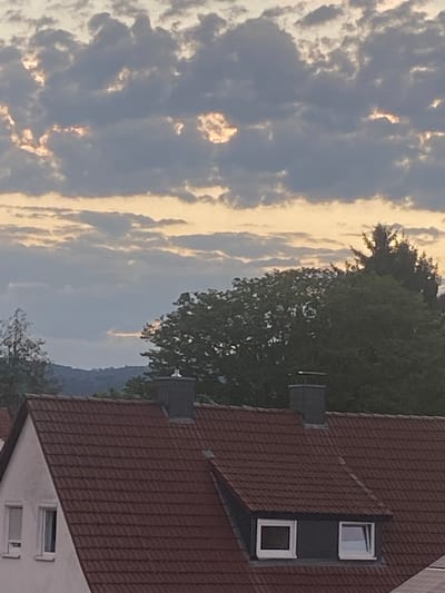 The image depicts a view of rooftops and trees against a dramatic cloudy sky. The clouds are a mix of dark grays and lighter grays with patches of the sky illuminated by the sun, creating a somewhat dramatic effect. A few tall trees, one with a distinctive conical shape, are visible behind the rooftops. The foreground shows the tiled roofs of houses, with chimneys and dormer windows. The style of the roofs and dormer windows suggests a residential area, typical of parts of Kassel, Germany. There are no people or animals visible in the frame. The overall impression is that of a late afternoon or early evening sky, possibly with some storm clouds gathering or breaking.