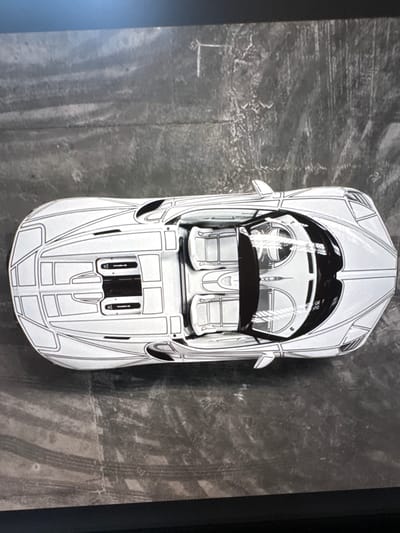 This is a top-down, medium shot of a white toy car with black accents on a dark, textured surface. The car is a convertible with visible interior details, including two seats and a dashboard. The exterior features a stylized design with bold black lines outlining panels and vents. The surface beneath the car appears to be concrete or asphalt, with visible tire marks and scuffs, suggesting an outdoor setting. The lighting is even, and there are no clear indications of time of day or weather. There is no visible text in the image. The overall impression is of a detailed toy car presented in a realistic environment.