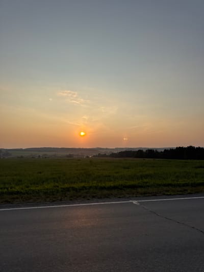 The image depicts a rural landscape in the vicinity of Большая Уса, Russia, during sunset. A paved road with a white line runs across the foreground, reflecting the fading sunlight. Beyond the road, a green field stretches towards a distant treeline. The setting sun, a bright orange orb, is visible in the hazy sky, casting a warm glow over the scene. The sky transitions from a pale blue at the top to a soft orange near the horizon, with a few wispy clouds present. In the distance, rolling hills and a dense forest can be seen, silhouetted against the colorful sky. There are no people or animals visible in the image. The overall impression is one of peaceful solitude in a natural setting.