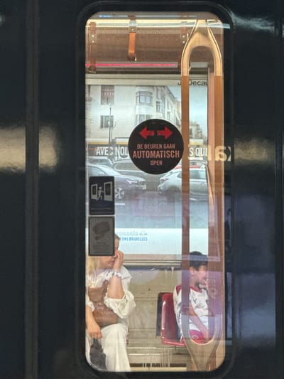 The image shows a view through a window, likely of a train or tram, looking outwards. Inside the vehicle, two people are visible. On the left, a young boy with dark hair is leaning his head against the window, looking outwards. He is wearing a white shirt with red and green accents. On the right, a woman with blonde hair is lying down, her head propped up by her hand. She is wearing a white ruffled shirt and her legs are extended. She appears relaxed.
Outside the window, there are reflections and what appears to be an advertisement or signage. Visible text includes "DE DEUREN GAAN AUTOMATISCH OPEN" which translates to "THE DOORS OPEN AUTOMATICALLY" in Dutch, and "AVEC NOUS" which is French for "WITH US". Another piece of text reads "BRUXELLES" (Brussels). There are also pictograms, one appearing to indicate accessible entry and another with a wheelchair symbol. The scene suggests a public transportation setting in Brussels, Belgium. The lighting and reflections indicate it is daytime.