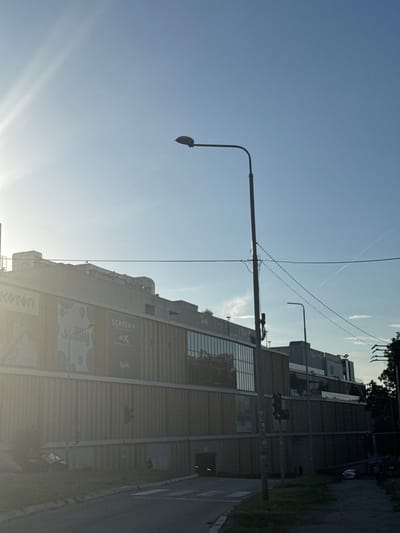 The image shows a wide, low-angle shot of a modern, multi-story commercial building with a beige, corrugated facade under a bright, hazy sky. The building features large windows on the upper levels and prominent ventilation systems on the roof. Several signs are visible on the building's facade, including "Koton" on the left and "ScreenX 4DX" on the right. A street lamp with a curved arm is positioned in the foreground, to the right of the frame's center. Another street lamp is visible further back on the right side of the building. A traffic light on a pole to the right of the central street lamp displays a green light. The foreground shows a paved road and a grassy area. In the distance, a white car is visible on the road to the right. The image appears to be taken in the city of Cukarica Urban Municipality, Serbia, given the presence of modern urban infrastructure and commercial establishments.