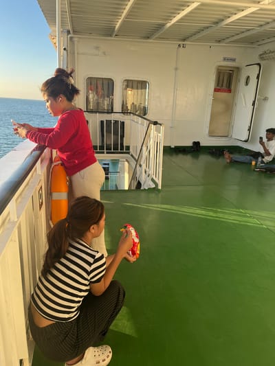 The image depicts a ferry deck with several individuals. In the foreground, two young women are present. One woman, wearing a red long-sleeved shirt and light-colored pants, leans on a railing, looking at her phone. She has her hair tied up in a bun and is wearing earbuds. An orange life preserver hangs on the railing beside her. The second woman, crouched down and wearing a black and white striped shirt and striped pants, holds an open bag of chips and appears to be eating from it. She is wearing white clogs. In the background, a young man in a white t-shirt and blue jeans is sitting on the green deck, looking at his phone. Several pairs of shoes are scattered on the deck near him, and a bottle of water is next to him. A white door with a sign reading "ANJUNGAN" above it is open. The deck is painted green, and the exterior of the ferry is white. The sea is visible in the background under a clear blue sky. The lighting suggests it is daytime, possibly late afternoon due to the warm light.