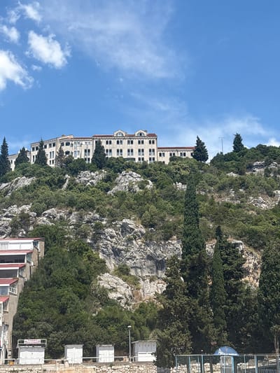The image shows a hillside in Neum, Bosnia and Herzegovina. The hillside is covered in trees and rock outcroppings. A large building is situated on the hillside. The sky is blue with a few clouds. Some commercial signage is visible on the right side of the image.