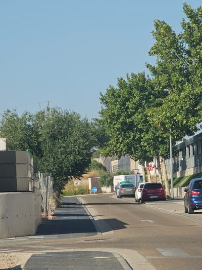 The image shows a street scene in Cuarte de Huerva, Spain. In the foreground, a paved walkway runs parallel to the street, with a concrete barrier on the left. Several cars are parked along the right side of the street, including a blue hatchback, a maroon sedan, and a white hatchback. A white van is also visible further down the street. Trees with green foliage line the right side of the street, and a modern building with a gray facade is in the background. The sky is clear and blue. A blue directional arrow sign is visible on the right side of the road.