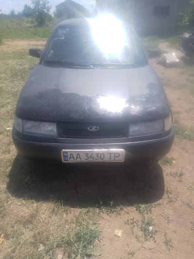 A dark-colored car is parked on a dirt and grass surface. The car is facing forward and is the main subject of the image. The license plate is visible and reads "AA 3430 TP" with "UA" on the left. The car's headlights and front grille are discernible, as is a logo on the grille. The sun is bright and reflecting off the car's windshield, creating a glare. In the background, there are trees, a building, and a cloudy sky. The environment appears to be rural or suburban, possibly in Inhulo-Kamianka, Ukraine.